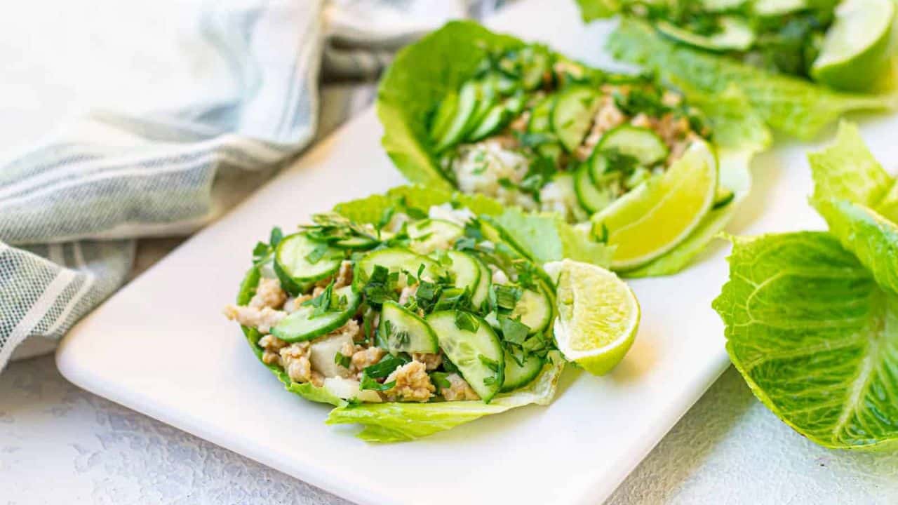 Lettuce wraps filled with sliced cucumbers, herbs, and ground meat on a white platter, served with lime wedges.