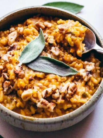 A bowl of pumpkin risotto topped with walnuts and a sage leaf, next to a pumpkin and sage leaves on a white surface.