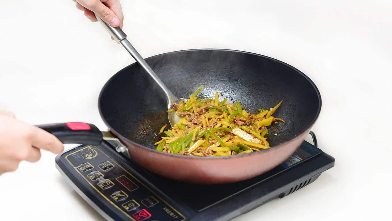 A person is using a large spoon to stir-fry sliced yellow and green vegetables in a wok on an induction cooktop.