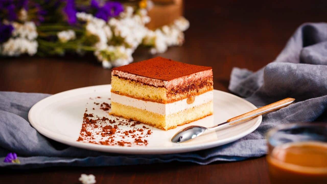 A slice of layered tiramisu cake with cocoa dusting, served on a white plate with a spoon, set on a dark table with a gray napkin and purple flowers in the background.