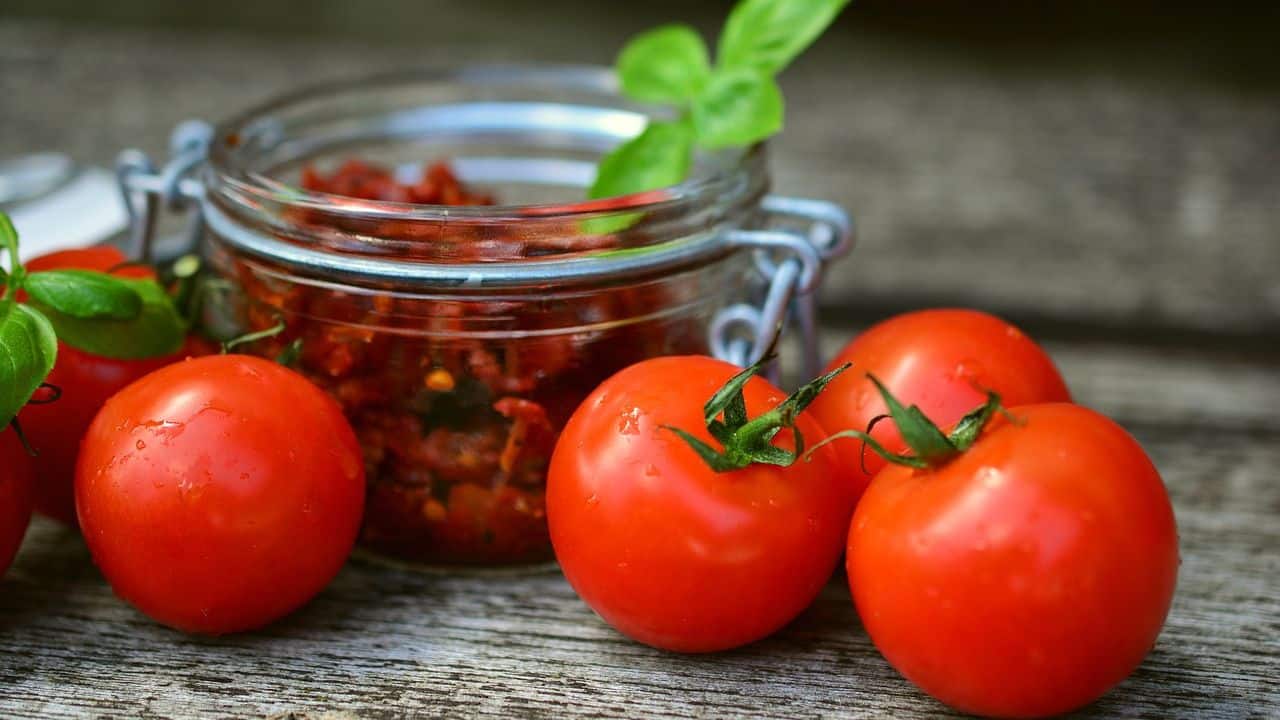 Jar of sun-dried tomatoes with fresh tomatoes and basil on a wooden surface.