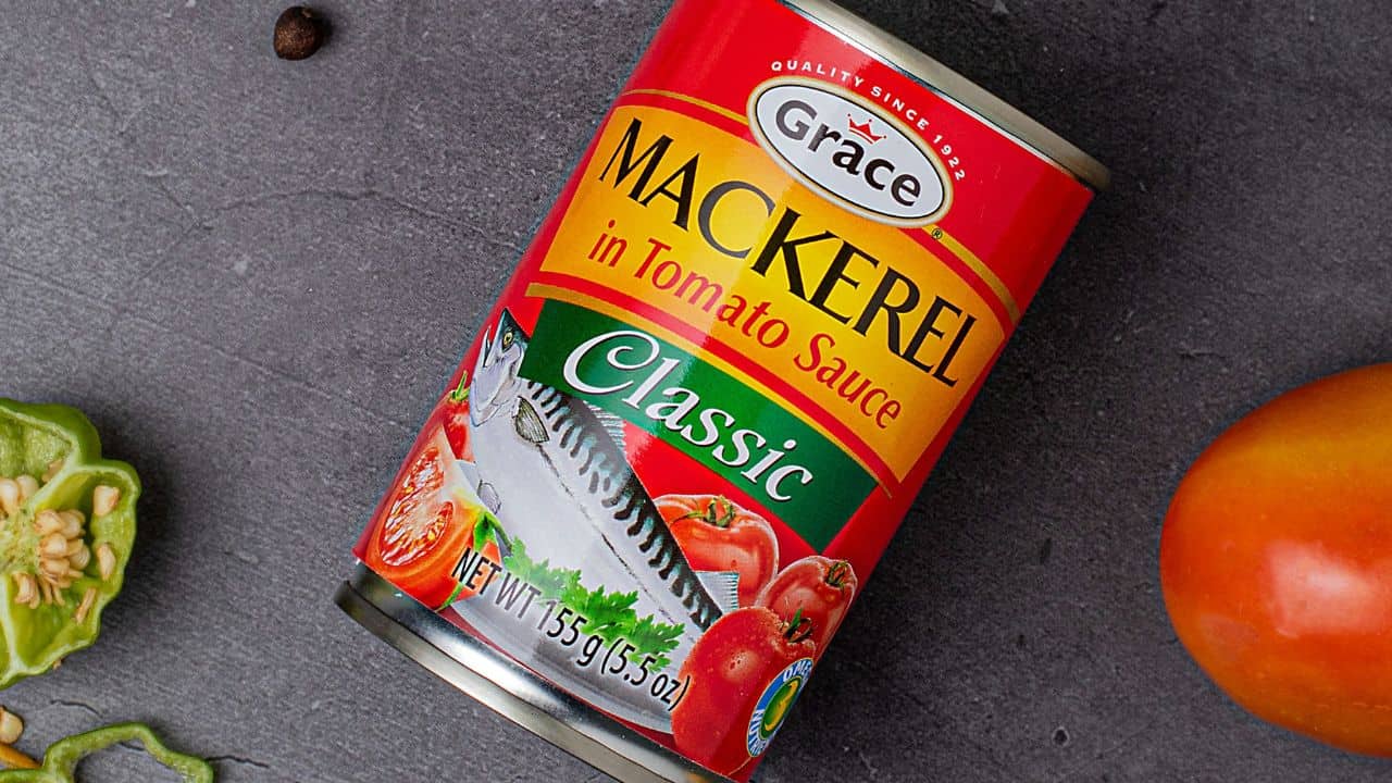 A can of Grace Mackerel in Tomato Sauce with a classic label design is placed on a dark surface, surrounded by a partially sliced green pepper and a whole tomato.