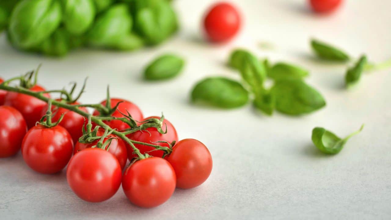 Cluster of red cherry tomatoes with scattered green basil leaves on a light surface.