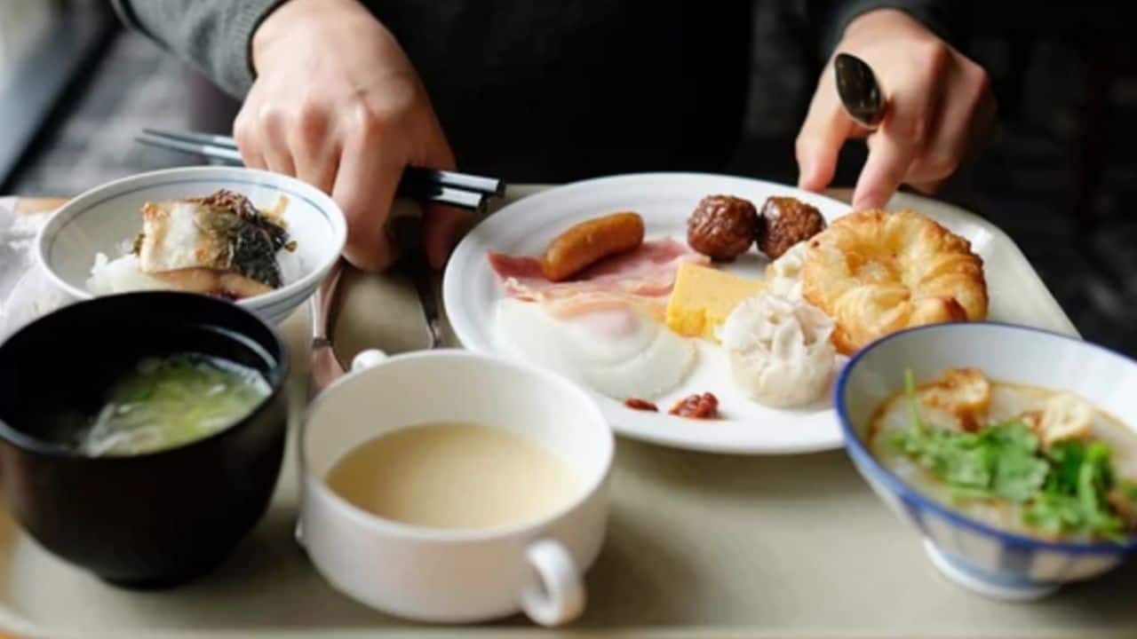 A person is sitting in front of a tray with a variety of foods, including eggs, ham, a croissant, meatballs, soup, and rice.