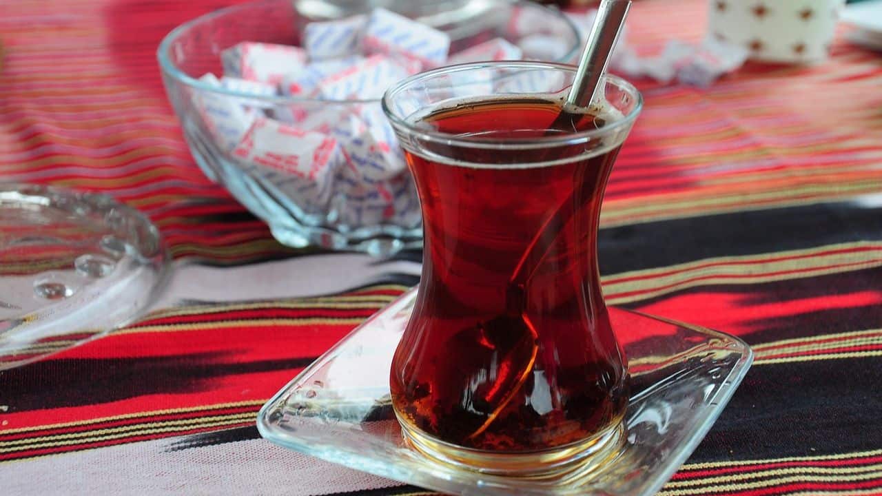 A glass of tea with a spoon on a square saucer sits on a striped tablecloth, with a bowl of sugar packets in the background.