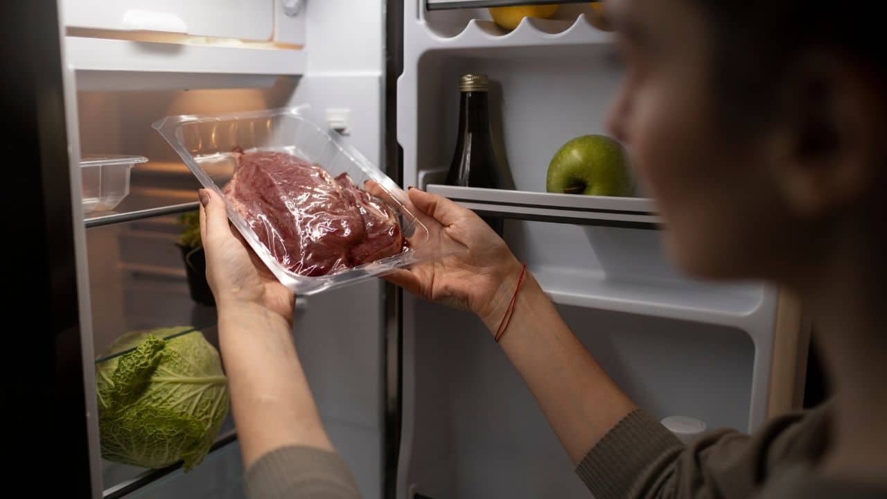 A person holds a plastic-wrapped piece of raw meat in front of an open refrigerator containing cabbage, an apple, and various items.