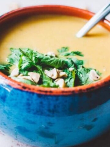 A blue bowl of soup garnished with cilantro and nuts, with a spoon resting inside.
