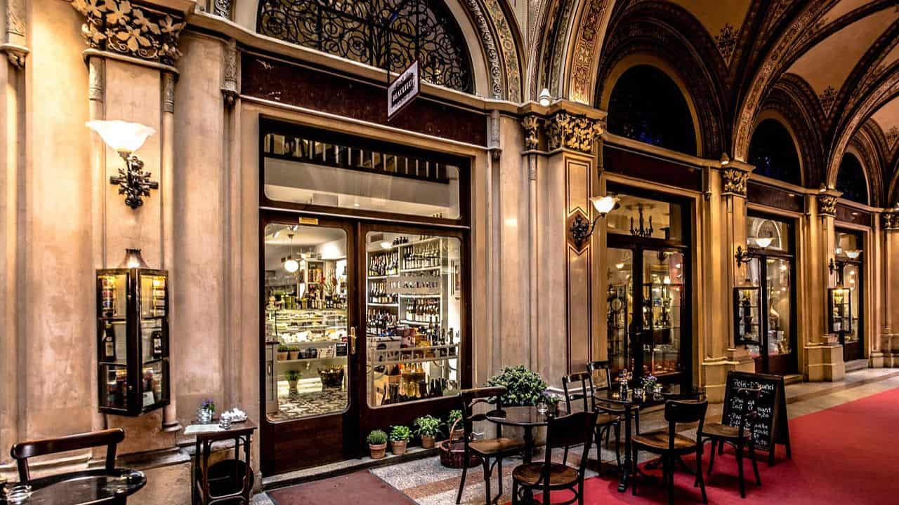 A cozy, dimly lit cafe with outdoor seating in an ornate, arched hallway. Glass doors reveal shelves of bottles and a display case inside. Small potted plants line the entrance.