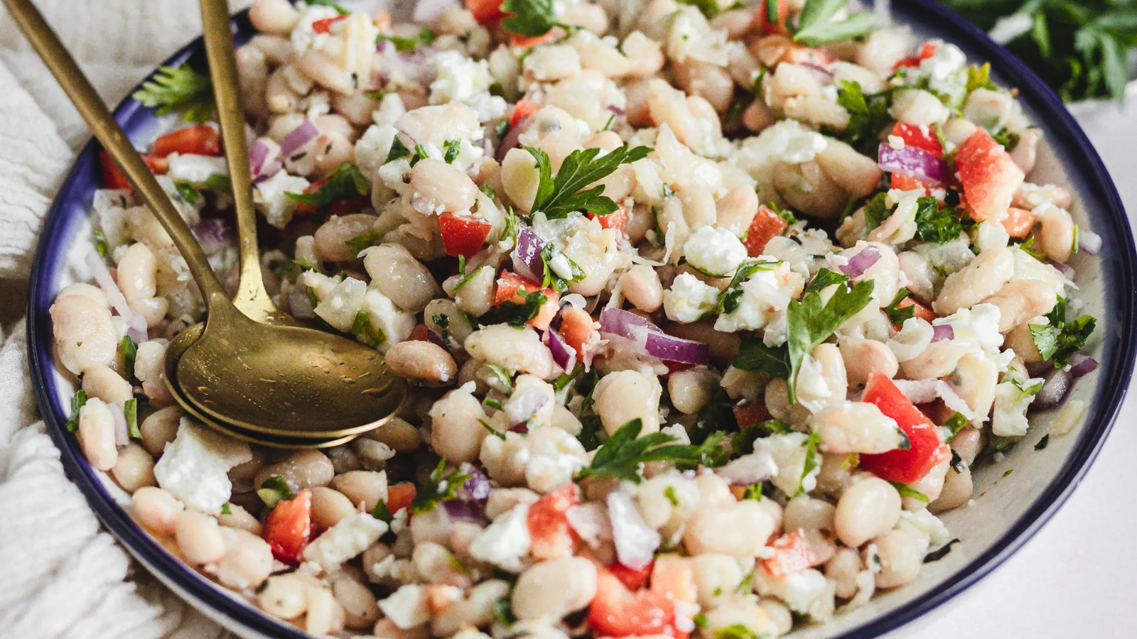 A bowl of white bean salad with chopped red peppers, onions, and parsley, garnished with cheese. Two gold spoons rest on the side.