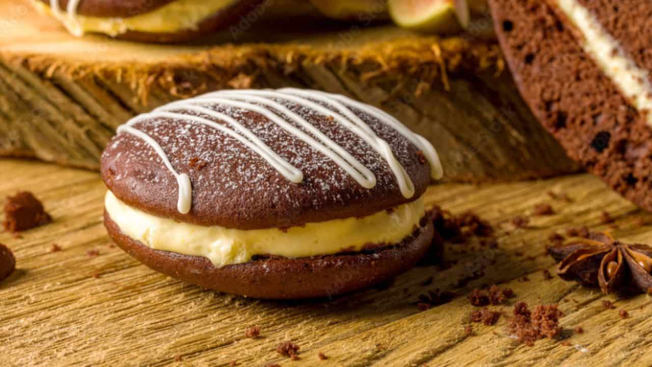 A chocolate whoopie pie with white frosting sits on a wooden surface, topped with a drizzle of white icing. Crumbs and anise stars are nearby.