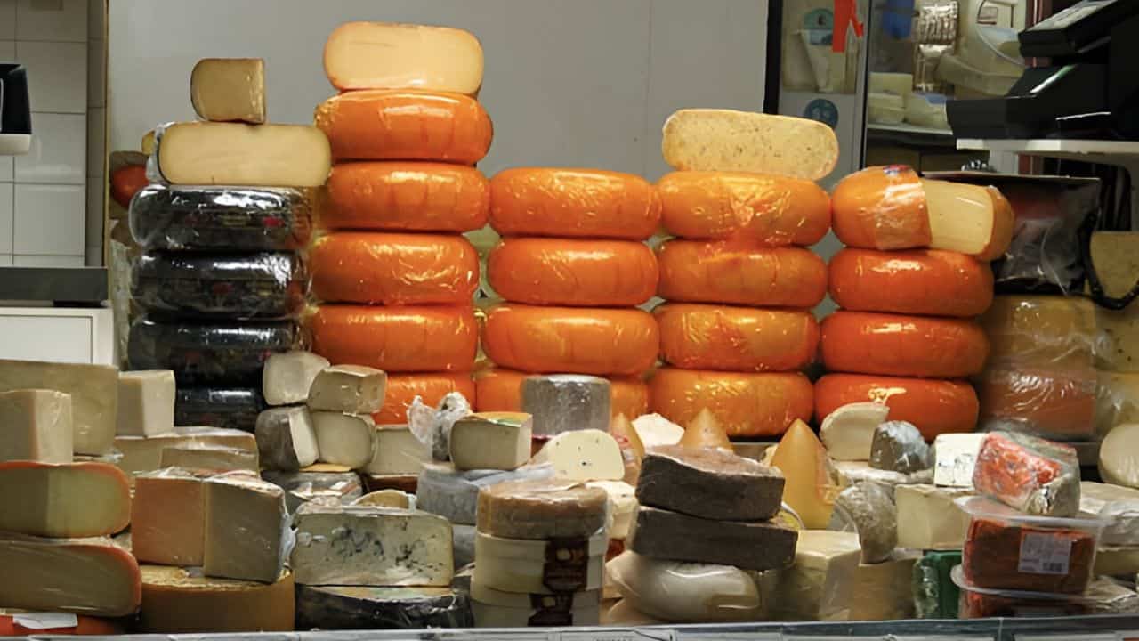 A display of various cheeses, including stacked red-waxed wheels and differently shaped and colored blocks, arranged on a countertop in a market setting.