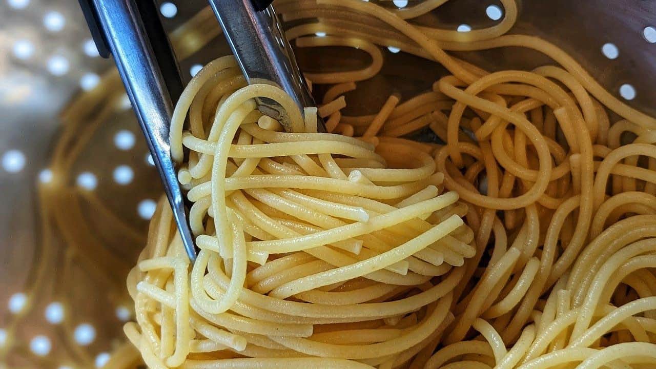 Close-up of cooked spaghetti being held with metal tongs over a perforated surface.
