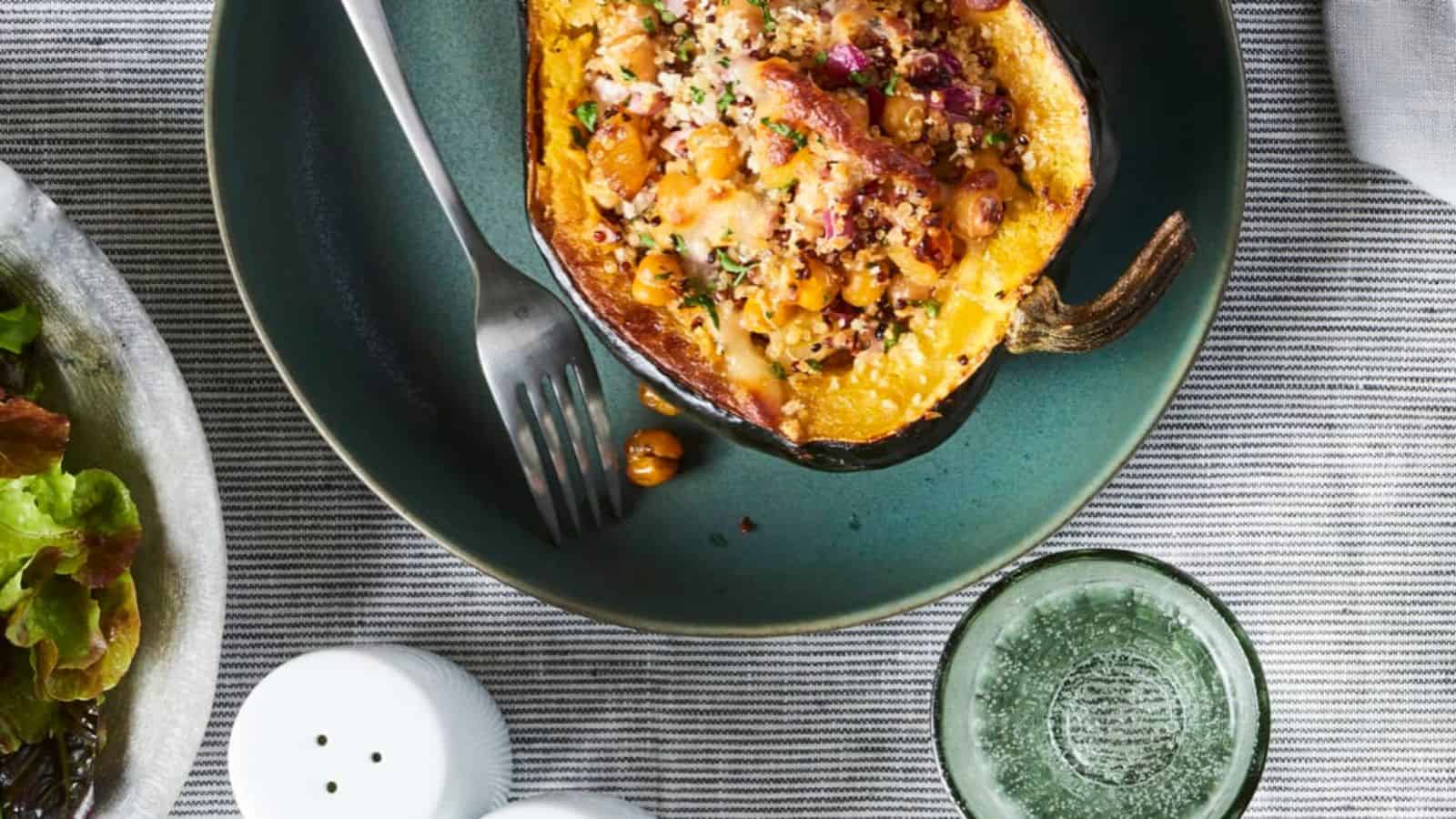 A stuffed acorn squash with quinoa and chickpeas on a plate next to a fork, with a glass and salt shakers nearby.