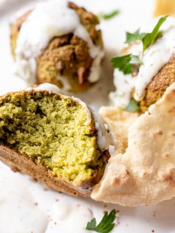 A plate of falafel with a sliced piece showing the interior, garnished with sauce and herbs, and accompanied by flatbread.