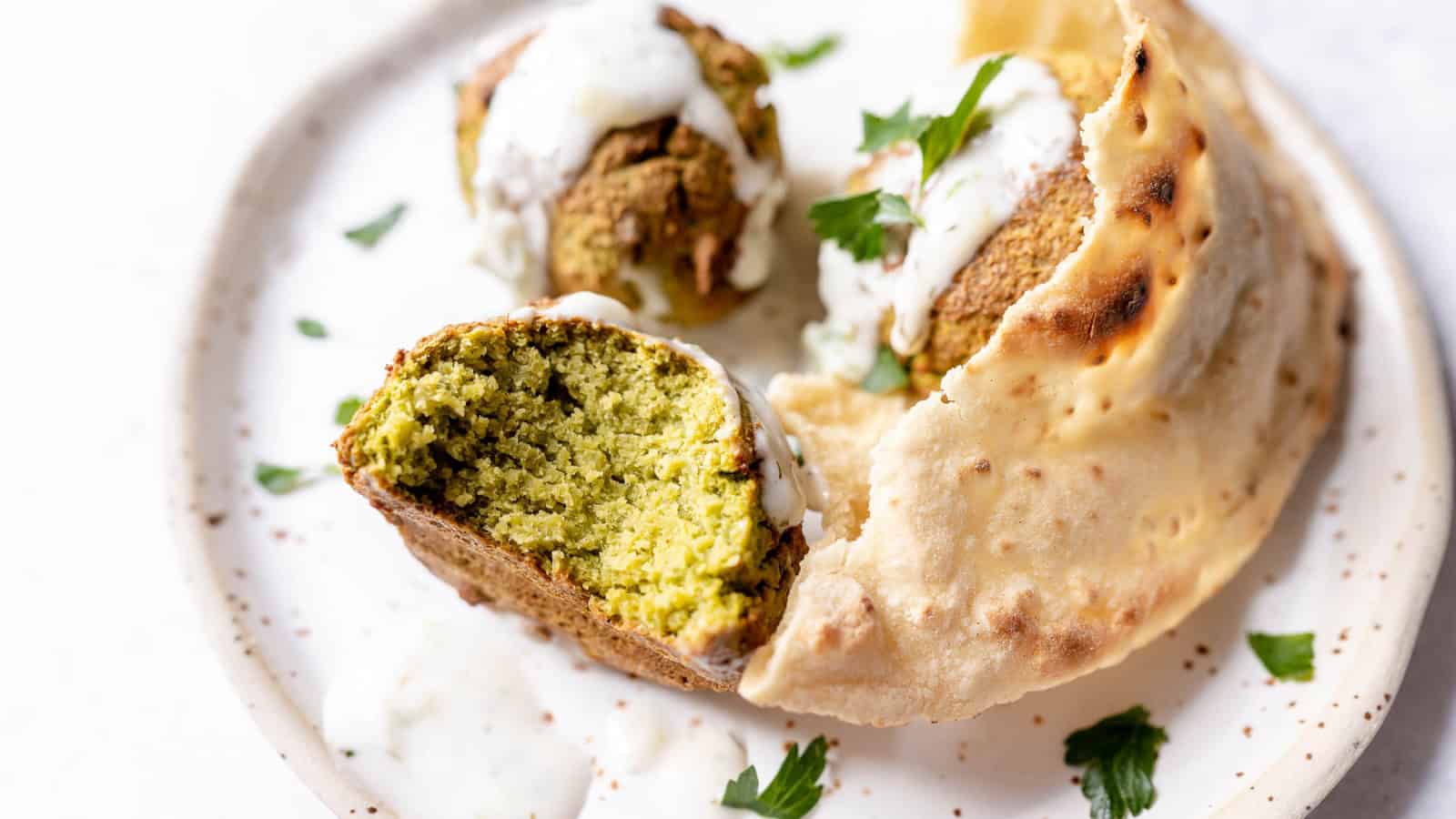 A plate of falafel with a sliced piece showing the interior, garnished with sauce and herbs, and accompanied by flatbread.