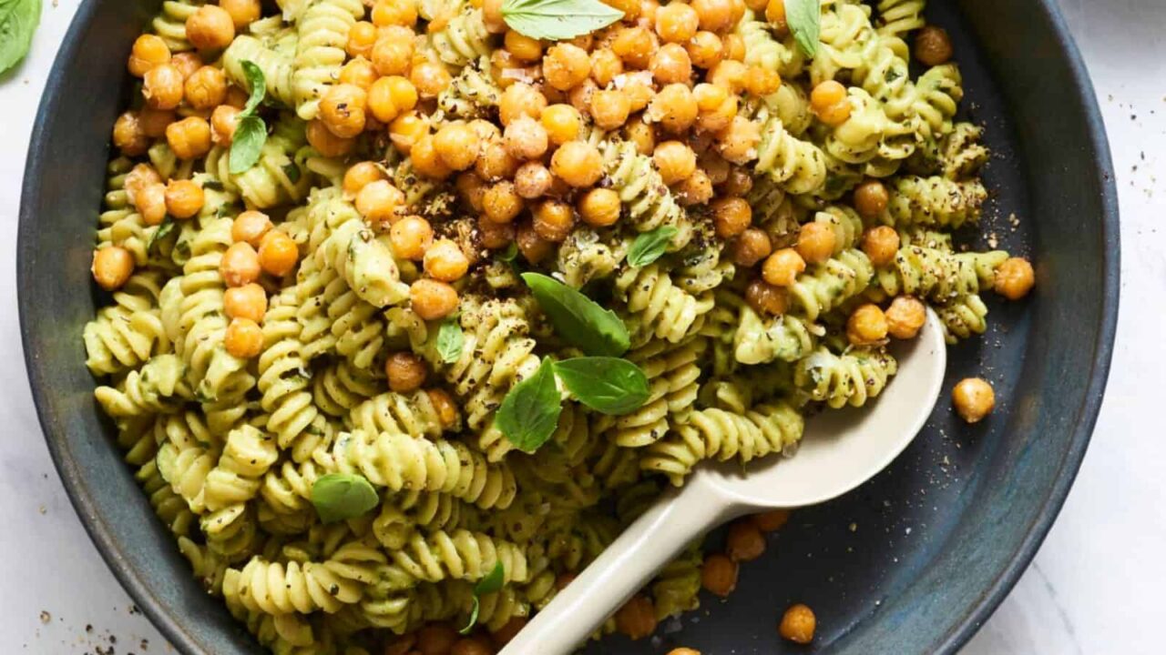 A plate of fusilli pasta with pesto sauce, topped with crispy chickpeas and basil leaves, with a spoon on the side.