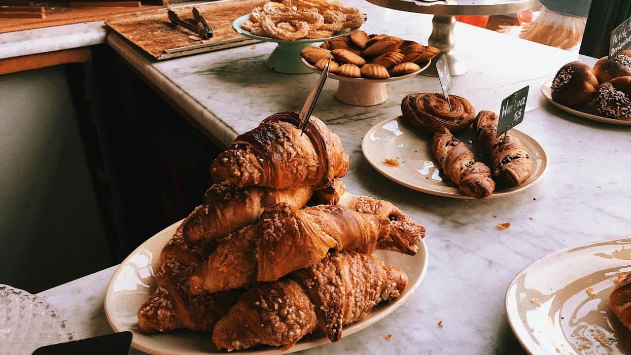 A marble countertop displays a variety of pastries, including croissants, cinnamon rolls, and round cookies on plates and a stand.