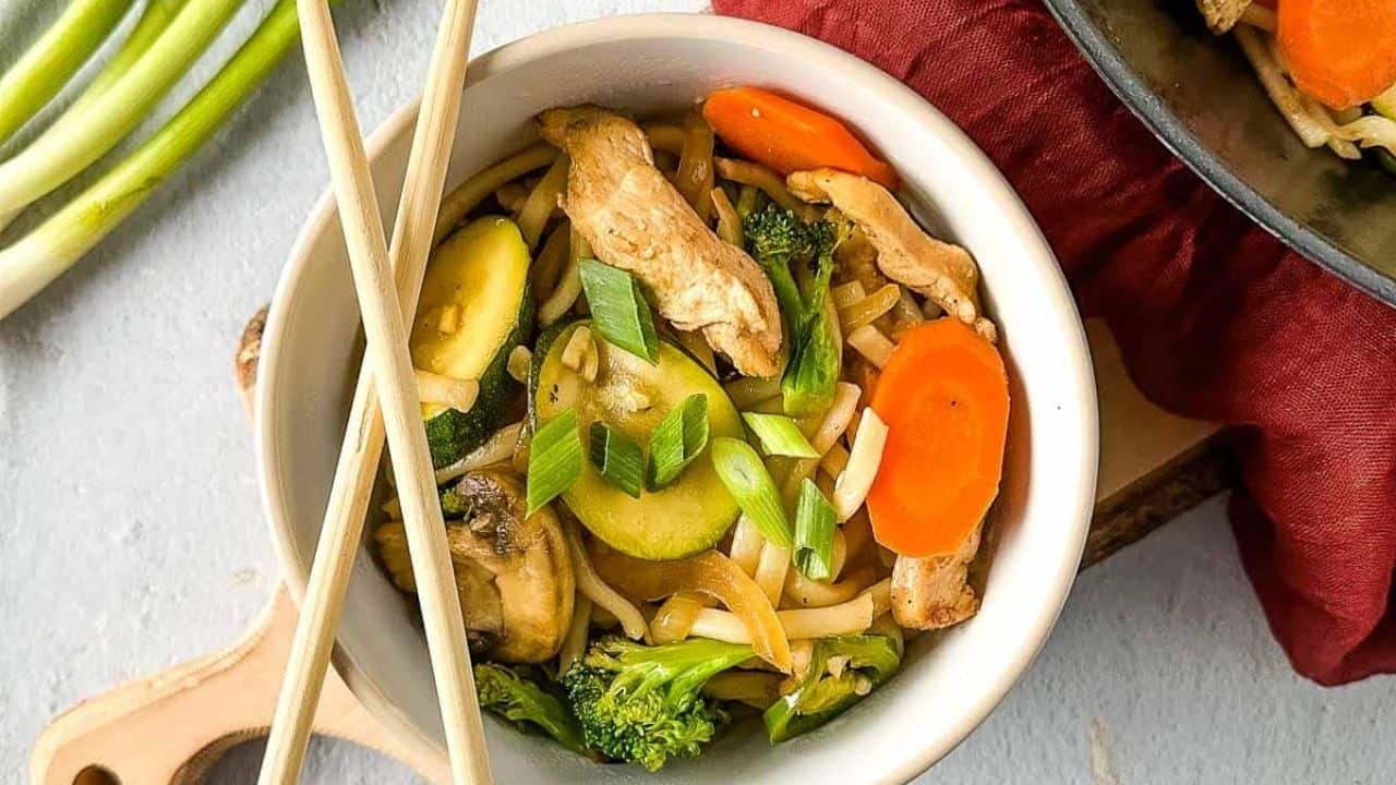 A bowl of stir-fried noodles with chicken, zucchini, carrots, broccoli, and green onions, accompanied by chopsticks.
