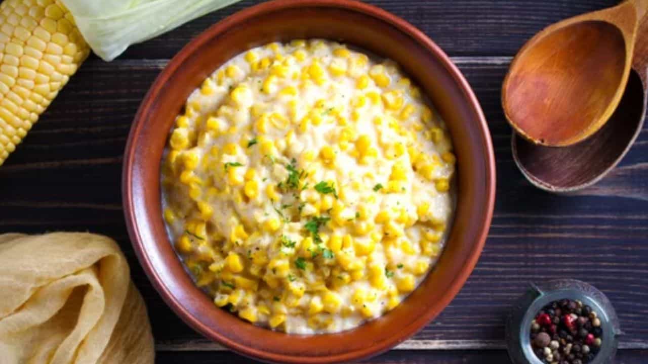 A bowl of creamy corn garnished with herbs on a wooden table, surrounded by a napkin, a wooden spoon, corn on the cob, and a small bowl of peppercorns.