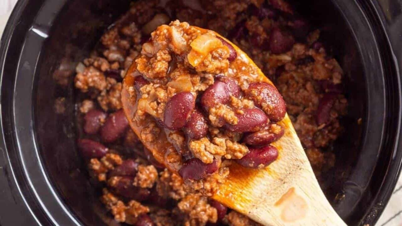 A wooden spoon scooping chili from a slow cooker, featuring ground meat, kidney beans, and diced onions in a tomato-based sauce.