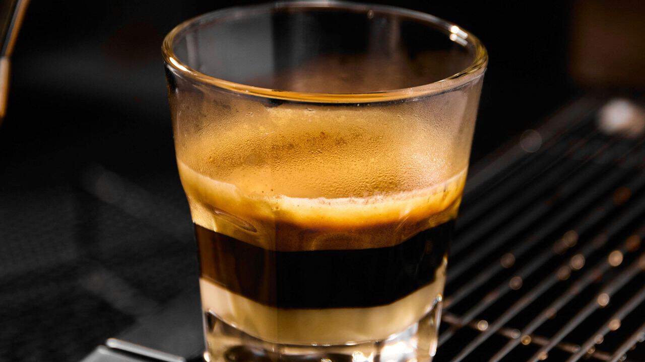 Close-up of a layered coffee drink in a clear glass, featuring distinct layers of espresso and milk, set against a dark background.
