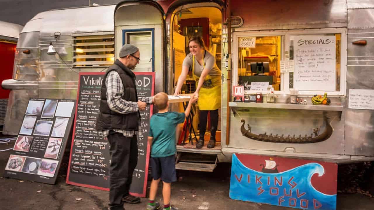 A customer with a child receives food from a vendor inside a retro food truck labeled "Viking Soul Food," with menu boards displayed outside.