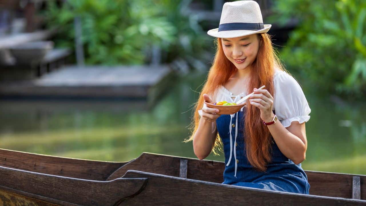A person in a hat sits in a boat on a calm body of water, enjoying a bowl of noodles with chopsticks.