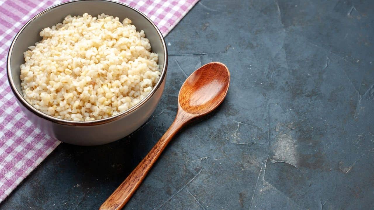 A bowl of cooked bulgur with a wooden spoon on a dark textured surface next to a pink and white checkered cloth.