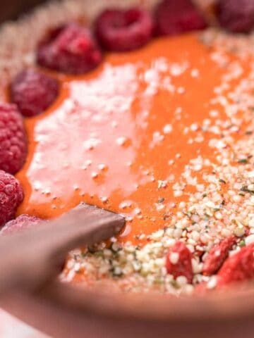A wooden bowl filled with a smoothie topped with raspberries, goji berries, and hemp seeds, with a wooden spoon resting inside.