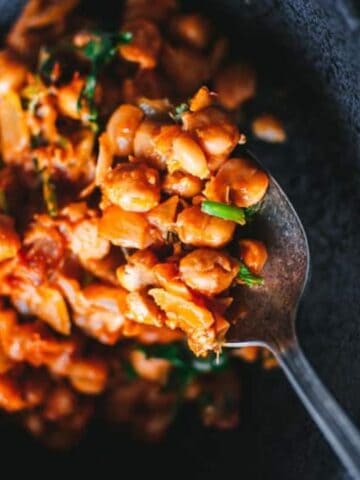 A spoon scoops a portion of baked beans mixed with herbs and onions from a dark bowl.