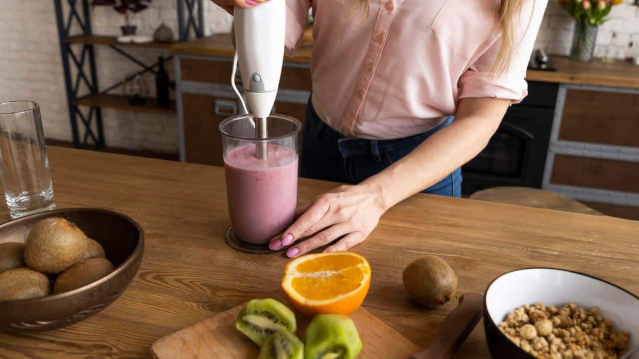 Person in a pink shirt using a hand blender to mix a pink smoothie in a glass, with kiwi, orange, and granola on the table.