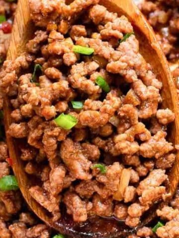 A wooden spoon scoops cooked, seasoned ground beef mixed with chopped green onions.