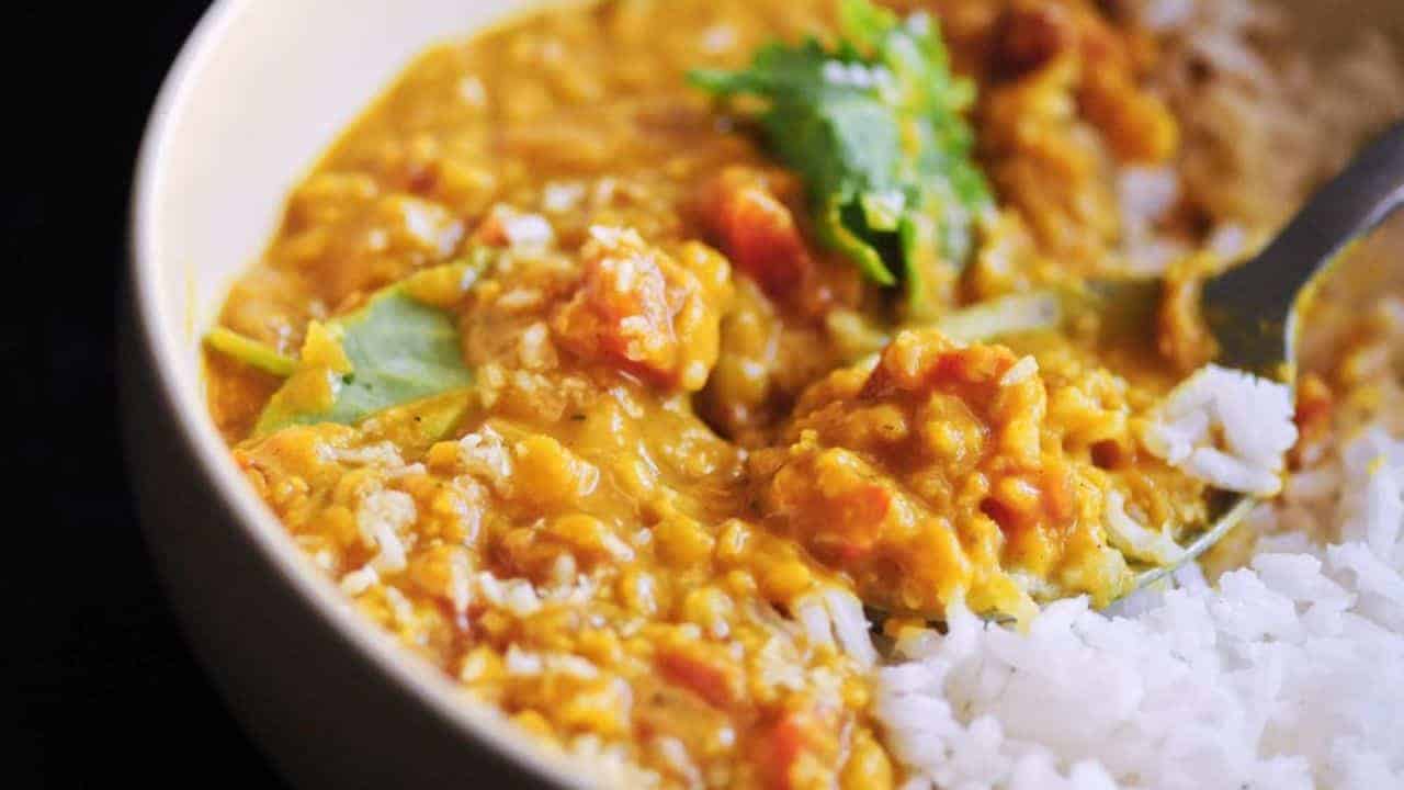 Close-up of a bowl of lentil curry with chopped tomatoes and leafy garnishes, served alongside white rice.