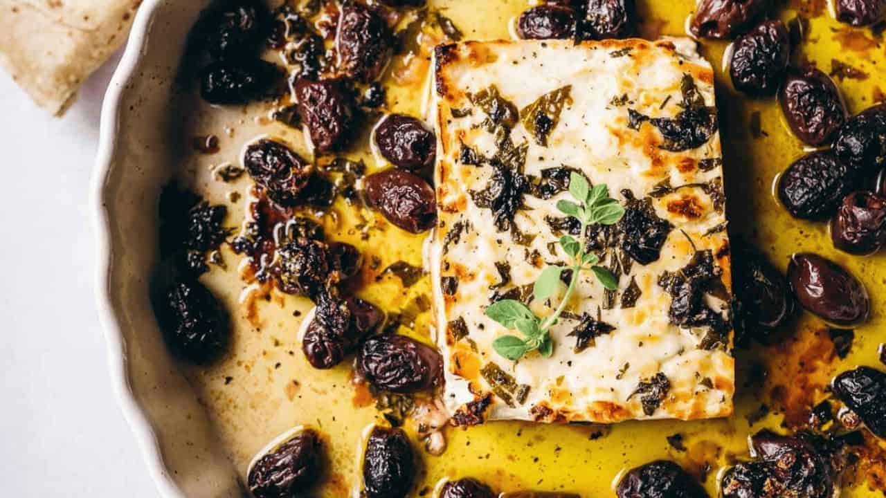 Baked feta cheese with herbs and black olives in a white dish with olive oil.