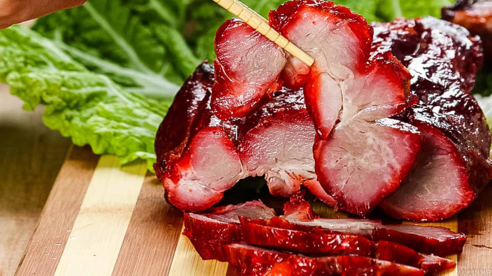 Close-up of sliced char siu pork on a wooden board, garnished with lettuce and being picked up with chopsticks.