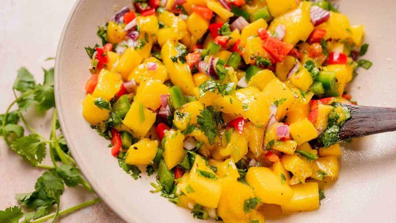 A bowl of mango salsa with diced mango, red onions, red and green peppers, and cilantro, mixed with a wooden spoon.