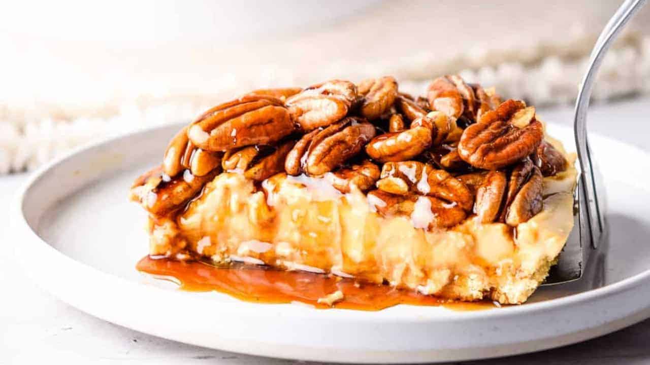 A slice of cheesecake topped with caramel and pecans sits on a white plate with a fork.