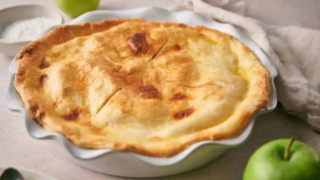A freshly baked golden-brown pie in a white pie dish, with a green apple and a small bowl of white cream nearby.
