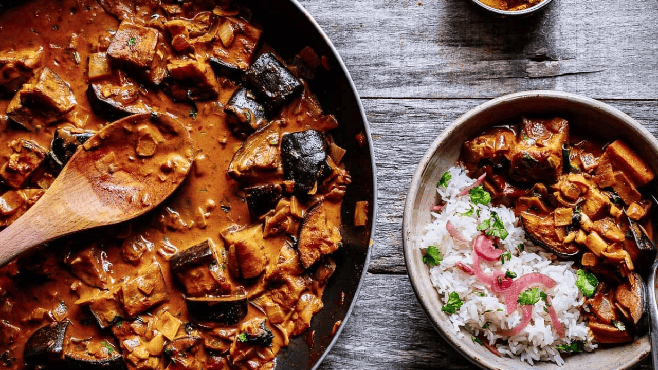 A pan of eggplant curry with a wooden spoon, and a bowl of rice garnished with cilantro and onion on a wooden table.