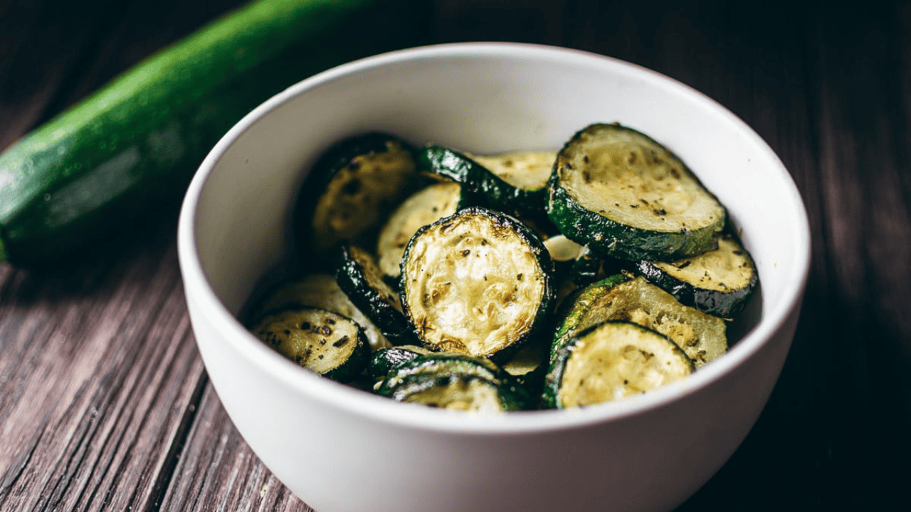 A white bowl filled with cooked, seasoned zucchini slices on a wooden surface.