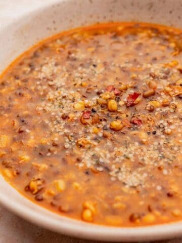 A bowl of hearty lentil soup with visible lentils and spices, garnished with seeds. A spoon rests inside the bowl on a light-colored surface.
