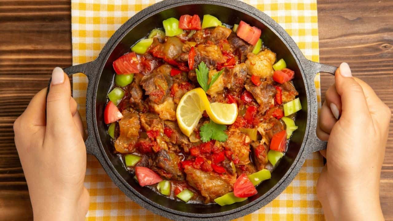 A cast iron pan filled with cooked vegetables, topped with lemon slices and cilantro, is held by two hands over a checkered cloth.