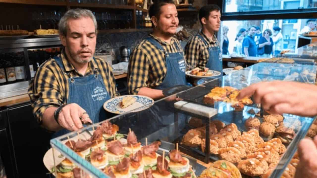 Three staff members in plaid shirts and aprons serve food from a bakery display filled with pastries and sandwiches.