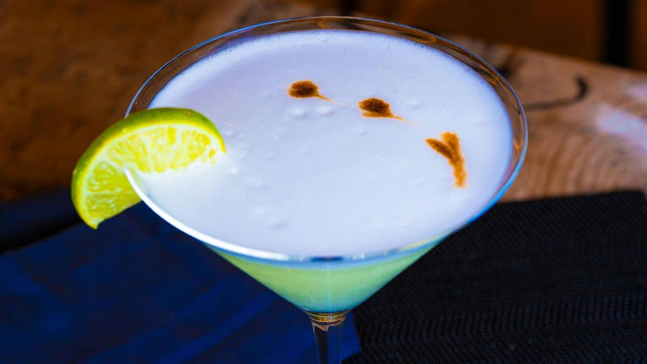 A cocktail in a martini glass garnished with a lime wedge, topped with frothy foam and three small brown drops.