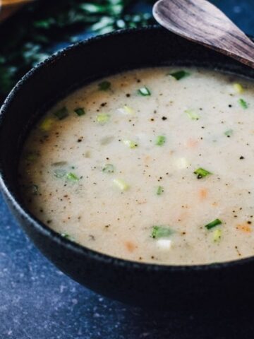 A bowl of creamy vegetable soup garnished with herbs, placed on a dark surface with a wooden spoon resting on the bowl's edge. Green onions and a small bowl of spices are nearby.