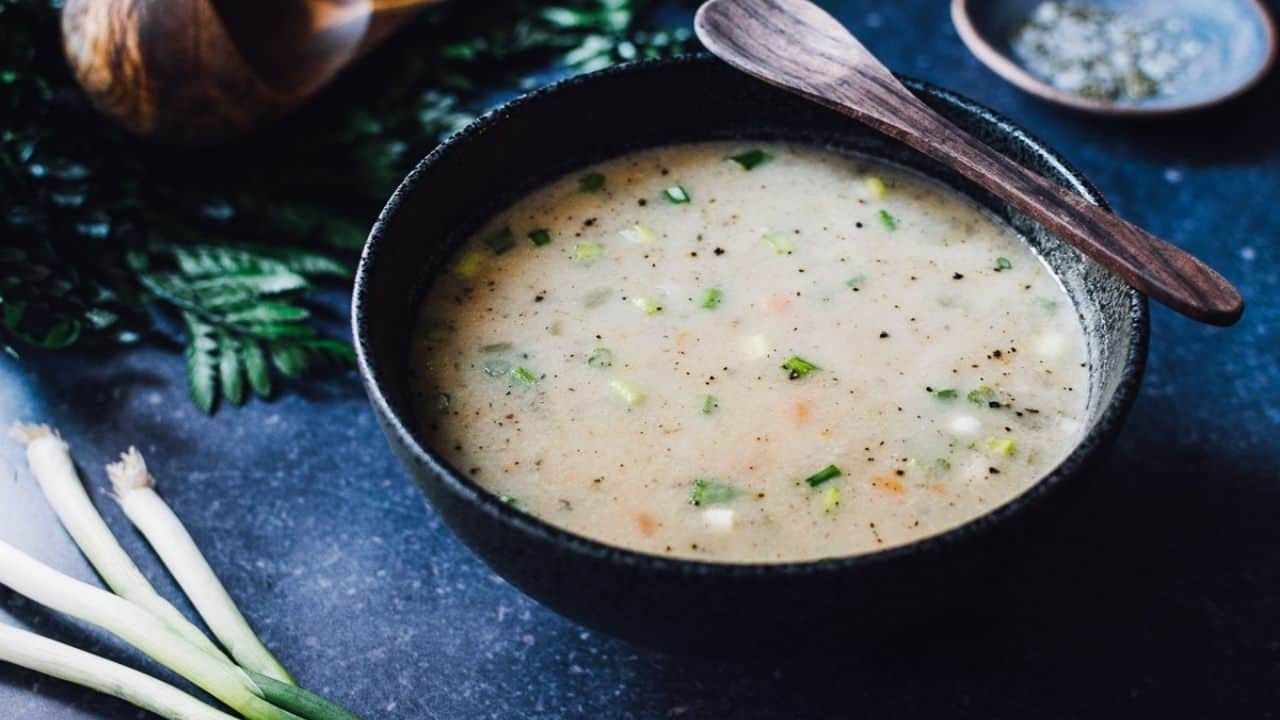 A bowl of creamy vegetable soup garnished with herbs, placed on a dark surface with a wooden spoon resting on the bowl's edge. Green onions and a small bowl of spices are nearby.