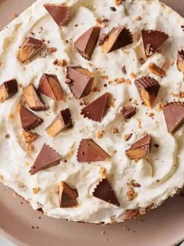 Overhead view of a round cake on a plate, topped with whipped cream and chunks of chocolate peanut butter cups. A knife and spoon are nearby on the table.