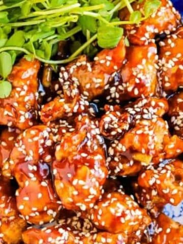 A plate of sesame-glazed fried chicken garnished with sesame seeds and fresh greens on a blue-patterned dish.