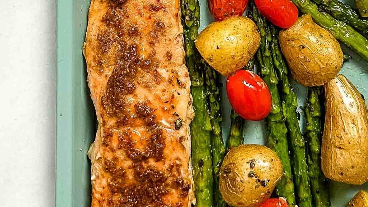 Grilled salmon with a brown glaze, accompanied by roasted baby potatoes, asparagus, and cherry tomatoes on a blue plate.