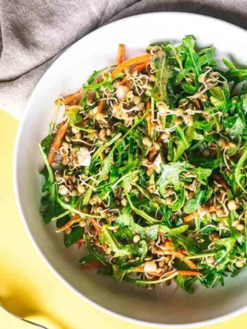A bowl of mixed greens, shredded carrots, and sprouts on a yellow surface, accompanied by a fork and napkin.